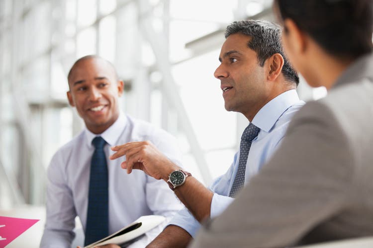 A diverse group of three professionals in business attire having an engaging discussion in a bright, modern office. A man in the center is speaking and gesturing, while a colleague in the background smiles and listens.