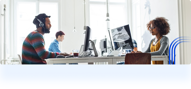 Three people working on laptops and monitors in a bright, open-concept coworking space. In the foreground, a man with headphones and a colorful sweater sits on the left, and a woman with glasses works on the right.