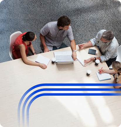 High-angle view of four professionals seated at a large, light-colored wooden conference table during a meeting. They are using laptops and taking notes. A stylized blue curved graphic is layered across the center of the table