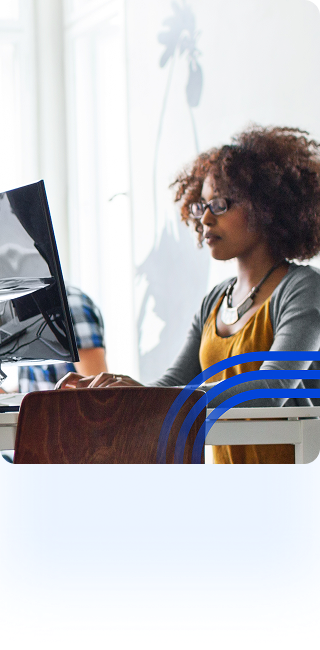 alt=&#34;Three people working on laptops and monitors in a bright, open-concept coworking space. In the foreground, a man with headphones and a colorful sweater sits on the left, and a woman with glasses works on the right.&#34;